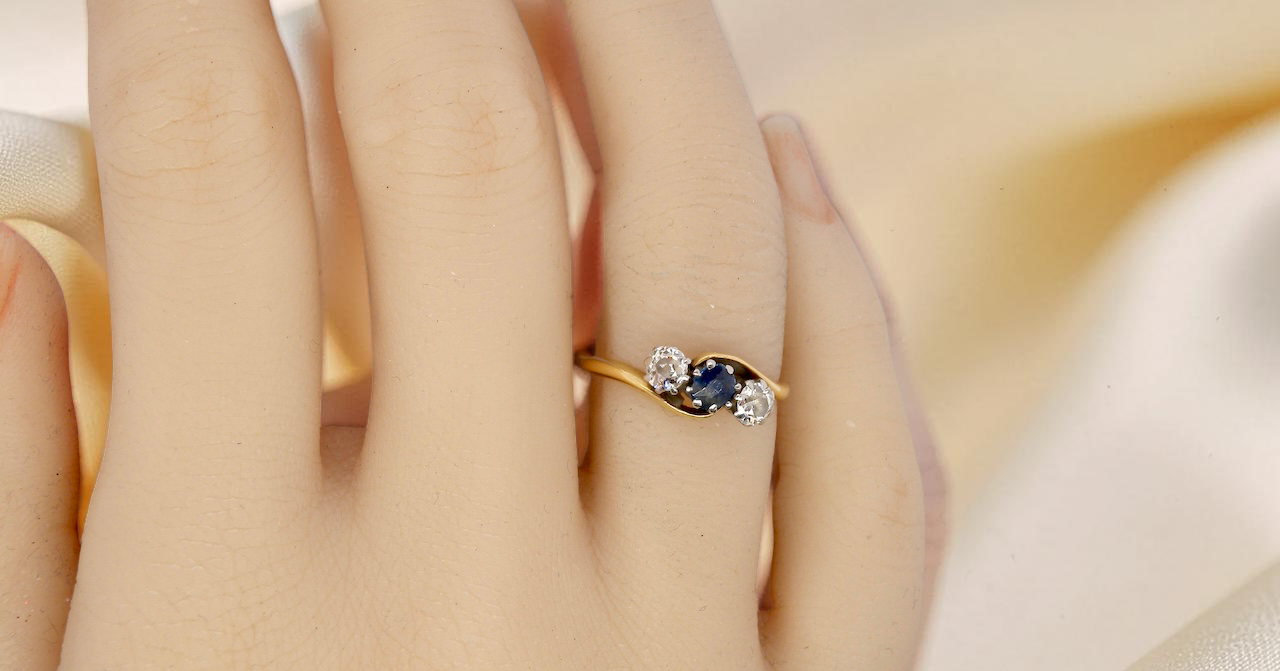 hand An 18 carat gold three stone cross over ring.
Centre stone a round sapphire 4mm diameter. Each side a brilliant cut white diamond 4mm diameter (0.25 carats estimated), colour i-k, clarity Si.