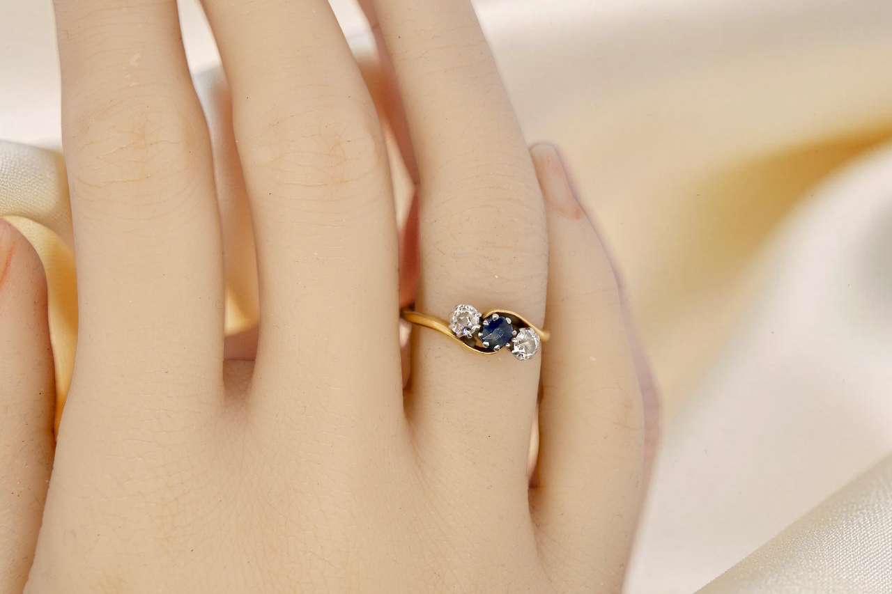 hand An 18 carat gold three stone cross over ring.
Centre stone a round sapphire 4mm diameter. Each side a brilliant cut white diamond 4mm diameter (0.25 carats estimated), colour i-k, clarity Si.