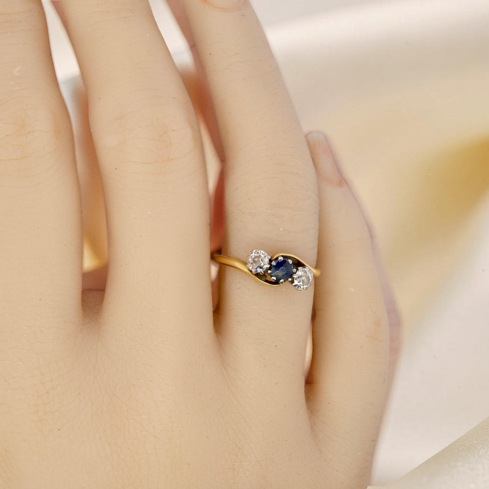 hand An 18 carat gold three stone cross over ring.
Centre stone a round sapphire 4mm diameter. Each side a brilliant cut white diamond 4mm diameter (0.25 carats estimated), colour i-k, clarity Si.