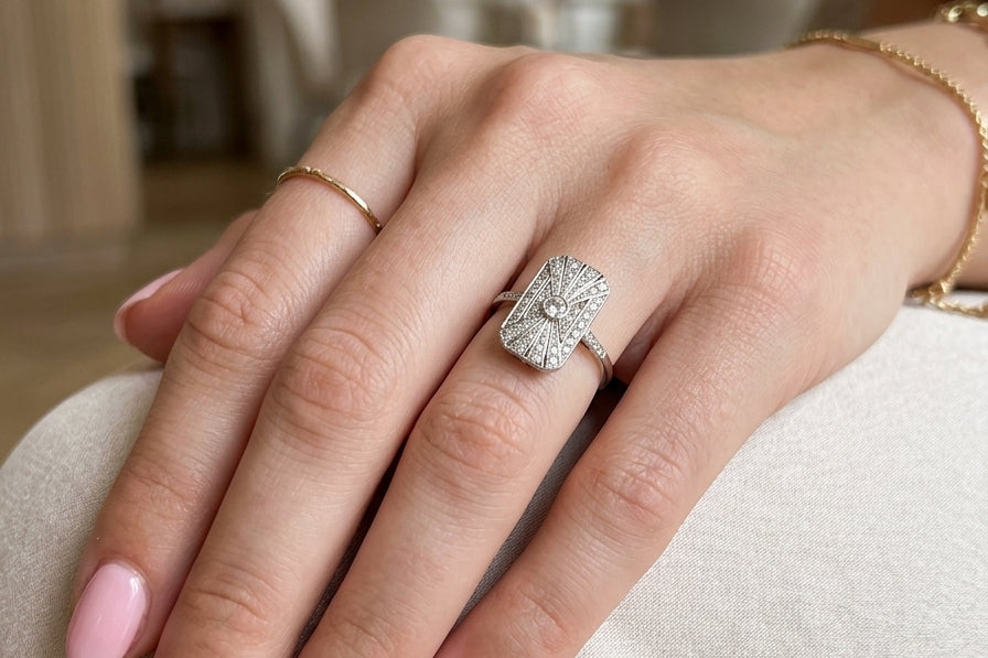 hand An Art Deco panel shaped cluster ring. The head measures 16x10mm, depth 3.4mm, and has two fan shaped designs that feature above and below a brilliant cut white diamond centre stone which measures 3.4mm (0.15 carats est)