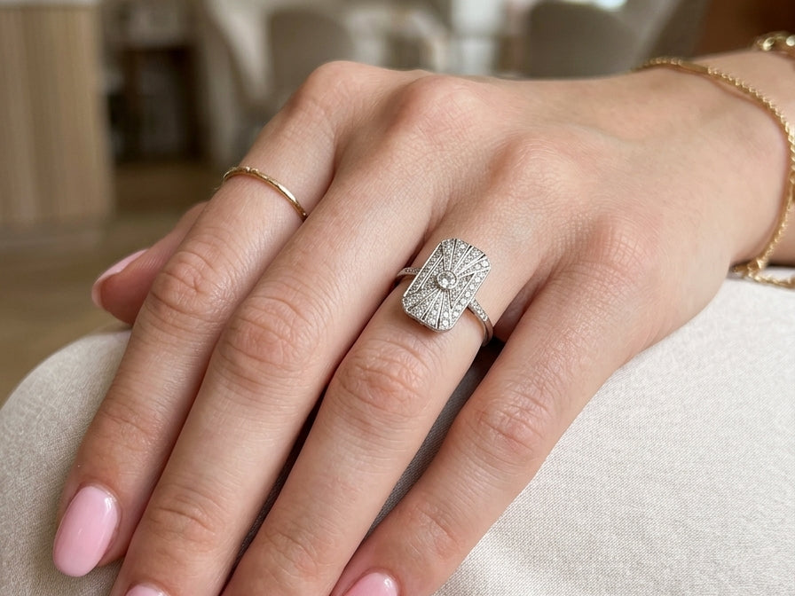 hand An Art Deco panel shaped cluster ring. The head measures 16x10mm, depth 3.4mm, and has two fan shaped designs that feature above and below a brilliant cut white diamond centre stone which measures 3.4mm (0.15 carats est)