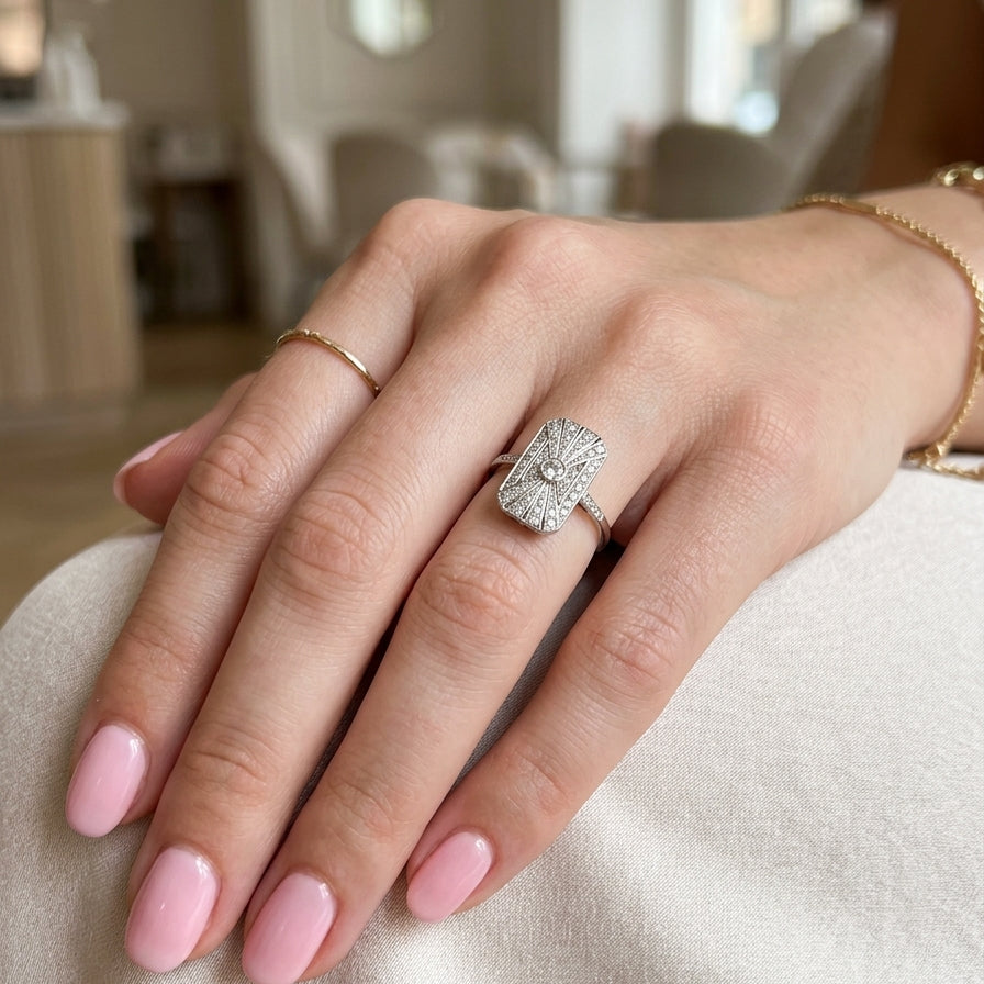 hand An Art Deco panel shaped cluster ring. The head measures 16x10mm, depth 3.4mm, and has two fan shaped designs that feature above and below a brilliant cut white diamond centre stone which measures 3.4mm (0.15 carats est)