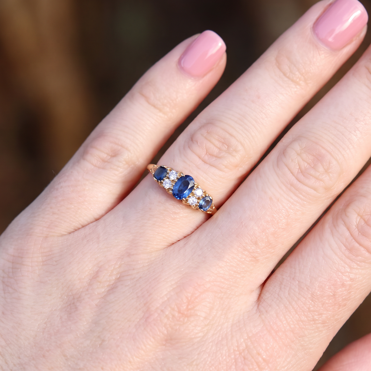 A close-up of a hand wearing a yellow gold ring with a central oval blue sapphire and smaller round blue and clear stones on the side, set against a blurred natural background.
