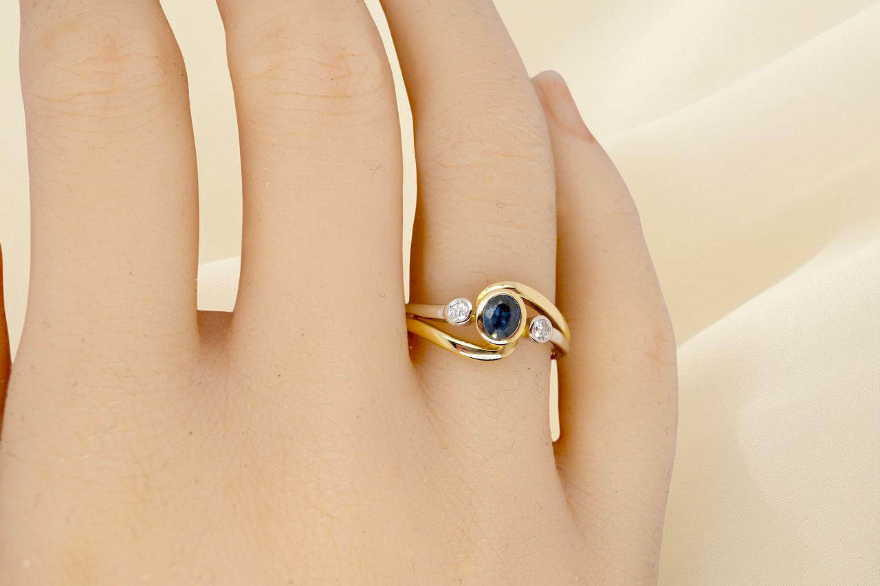 hand An elegant 18 carat gold three-stone ring featuring a rich, oval-cut dark blue sapphire at the centre, measuring 5 × 4mm, secured in a classic rim (bezel) setting.