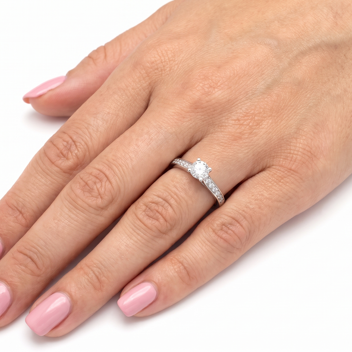 A close-up of a hand with pink manicured nails wearing a diamond engagement ring on the ring finger, set against a plain white background.