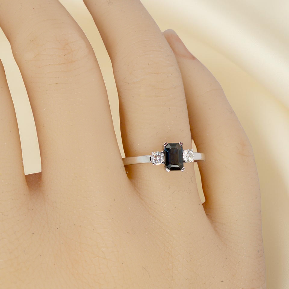 hand An elegant three-stone ring crafted in 18 carat white gold, featuring a striking rectangular dark blue sapphire at the centre, measuring 6 × 4 mm and weighing approximately 0.50 carats.