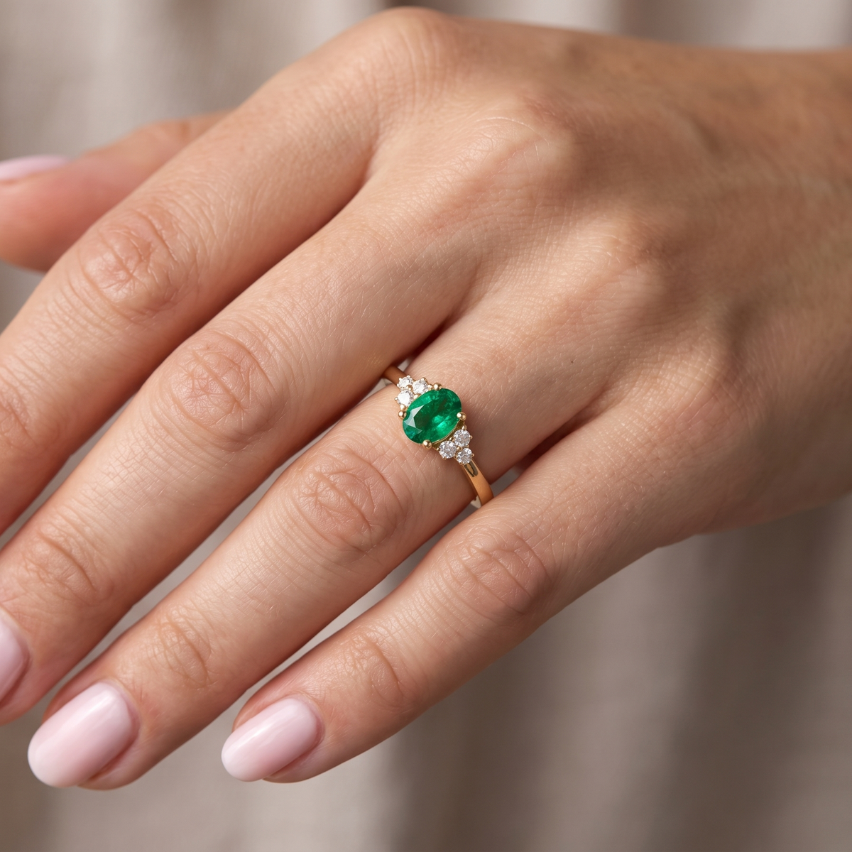 A close-up of a hand wearing a gold ring with a round green emerald-cut center stone and small clear accent stones on either side, set against a soft beige background.