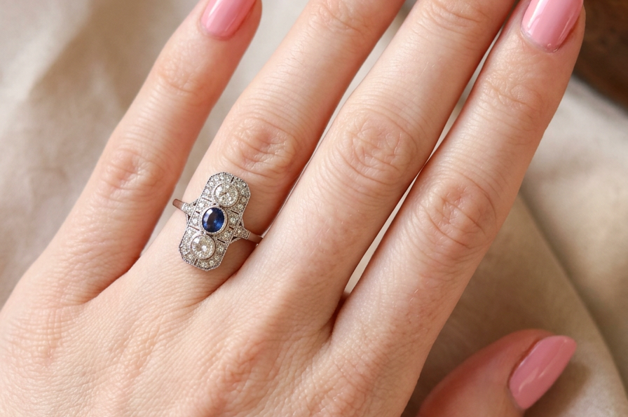 hand The plaque design has at its centre a mid to dark blue sapphire, 5x4mm (est 0.40 carats) mille grain set. Above and below to Old European Cut white diamonds. Each one estimated at 0.40 carats (4.8mm) Colour H-j, clarity Si.
