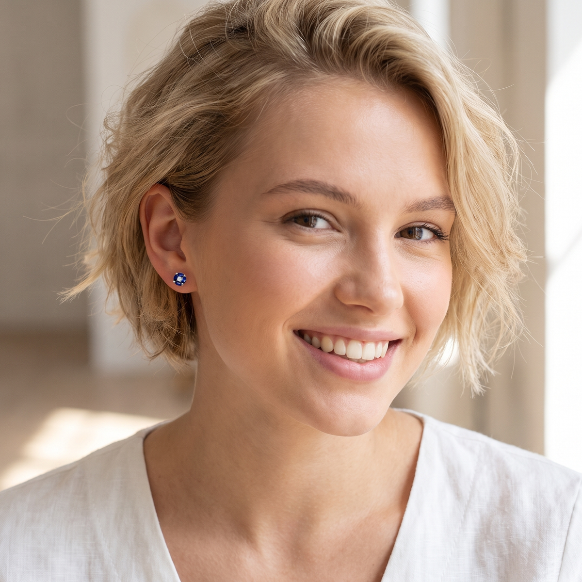A smiling blonde woman wearing a small blue stud earring, shown in a bright indoor setting with soft natural light.
