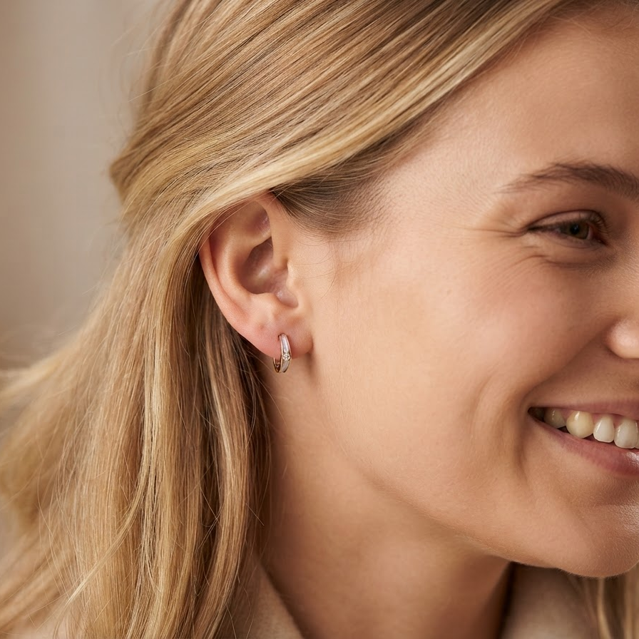 A close-up side profile of a smiling blonde woman wearing a small hoop earring with a pink stone and a tiny clear accent stone.