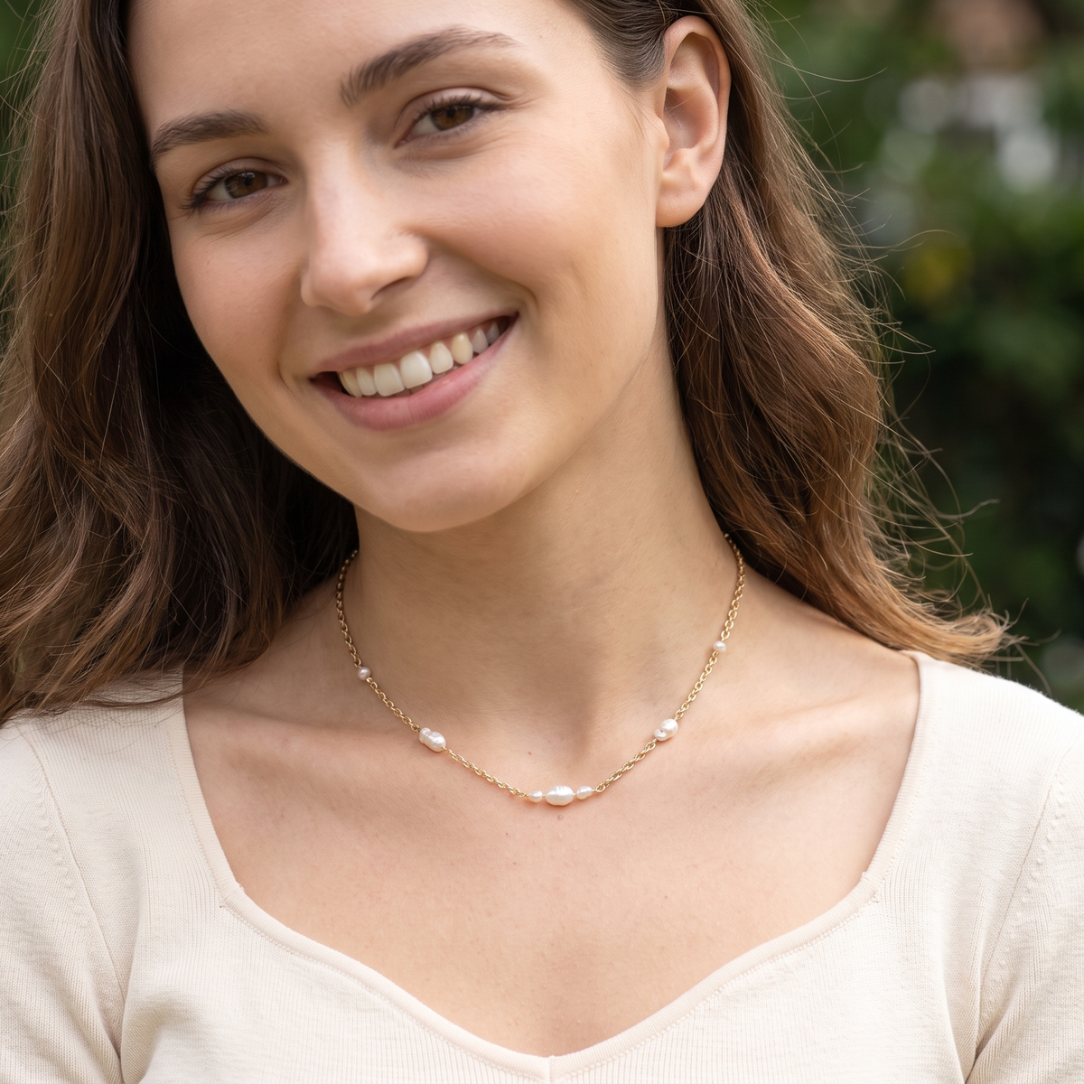 A smiling woman with long brown hair wearing a delicate gold chain necklace with small white pearl-like beads, photographed outdoors against a blurred green background.