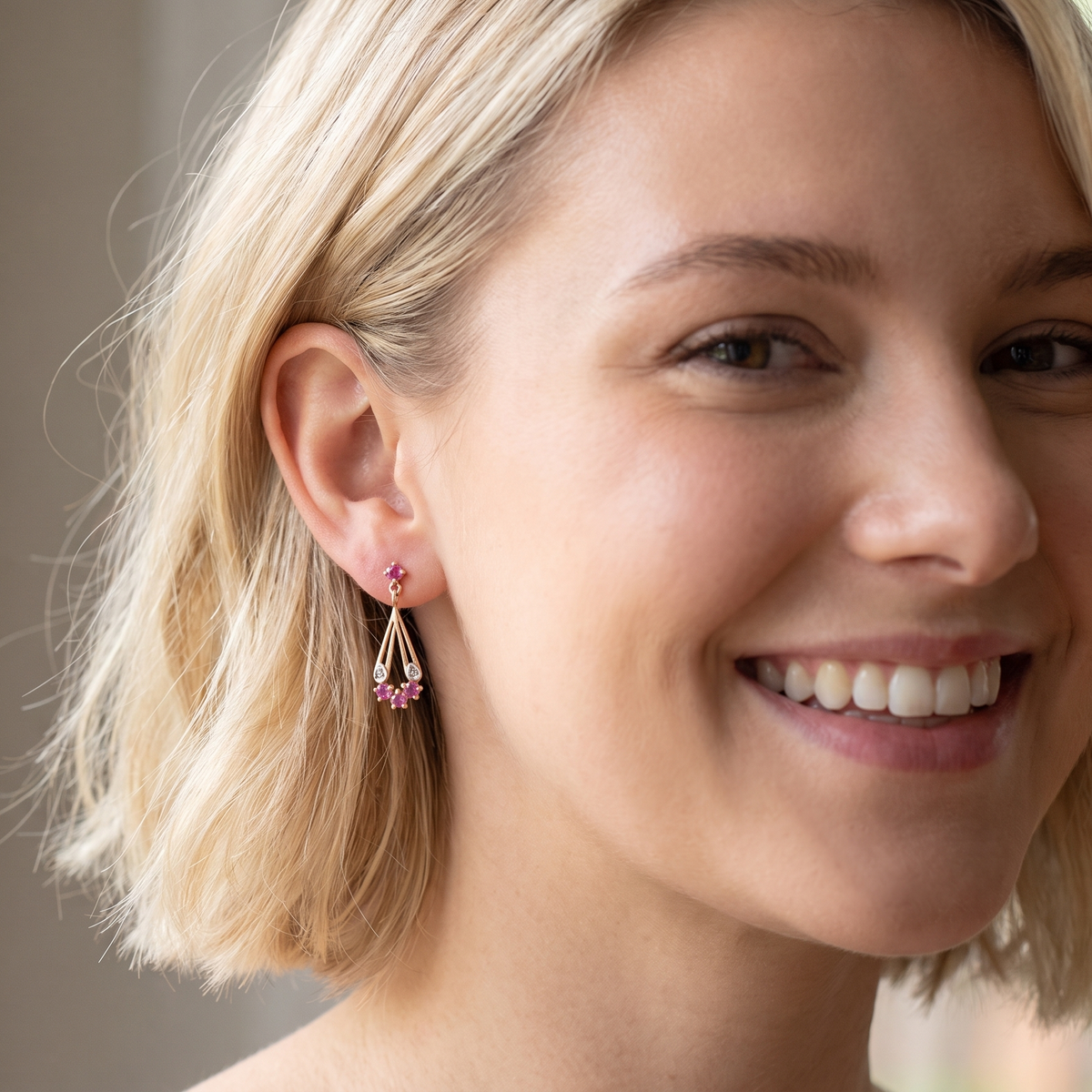 A close-up of a smiling blonde woman wearing a delicate gold dangle earring with small pink gemstones, set against a softly blurred indoor background.