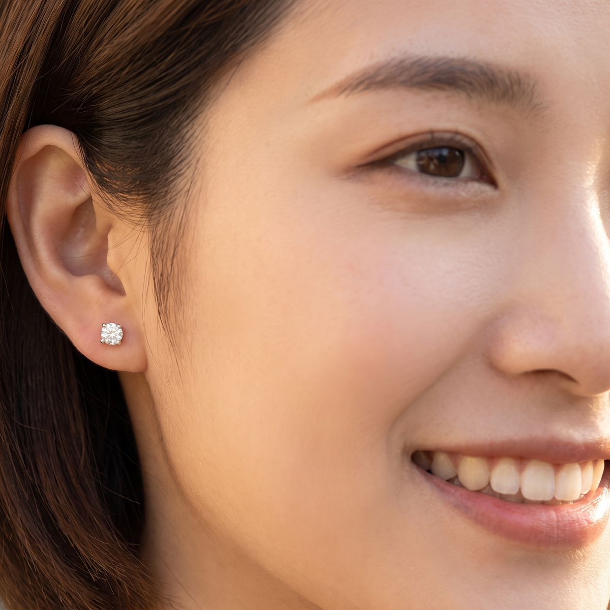 A close-up of a woman's ear wearing a small round clear stud earring, with her face partially visible in warm natural light.