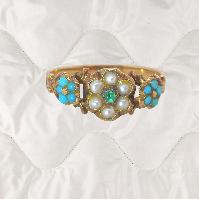 An antique Victorian gold ring with a floral design featuring white pearls, a central green emerald, and turquoise blue stones, displayed on a white quilted fabric background.