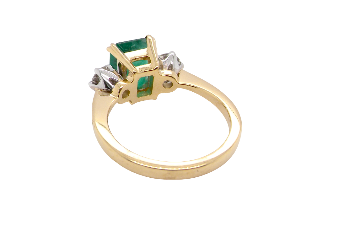 rear An exceptional three-stone ring featuring a vibrant step-cut emerald to the centre, measuring 8.6 x 7.0mm and weighing approximately 2.10 carats. The emerald displays a beautiful grass-green colour and is secured in a classic four-claw setting.