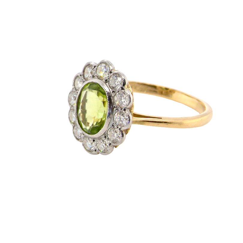 A yellow gold ring featuring an oval green peridot stone surrounded by a halo of small clear stones, set on a plain white background.