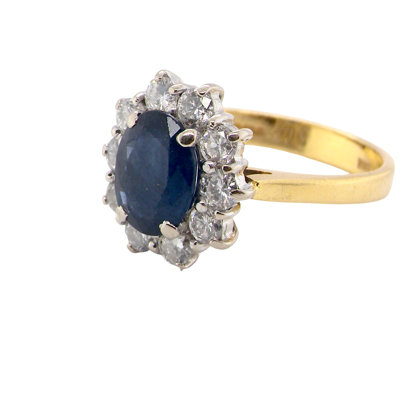 A yellow gold ring featuring an oval dark blue sapphire center stone surrounded by a halo of small clear stones, set on a plain white background.