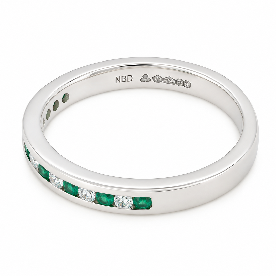 An eternity-style band ring featuring a channel setting of green emerald-cut gemstones and clear round stones, shown on a clean white background. The inside of the band is stamped with 'NBD'.