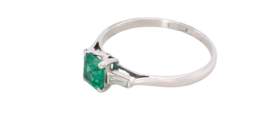 side An elegant and timeless platinum ring featuring a stunning step-cut emerald to the centre, measuring approximately 5.5 x 5.5mm and estimated at 0.80 carats. The emerald displays a vibrant, deep green colour with excellent presence.