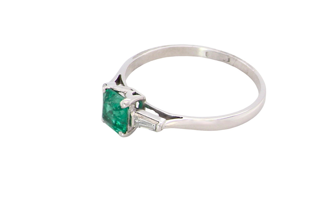 side An elegant and timeless platinum ring featuring a stunning step-cut emerald to the centre, measuring approximately 5.5 x 5.5mm and estimated at 0.80 carats. The emerald displays a vibrant, deep green colour with excellent presence.