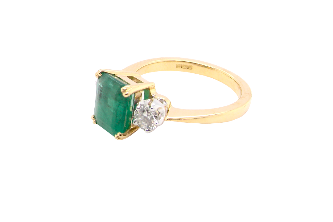 side An exceptional three-stone ring featuring a vibrant step-cut emerald to the centre, measuring 8.6 x 7.0mm and weighing approximately 2.10 carats. The emerald displays a beautiful grass-green colour and is secured in a classic four-claw setting.