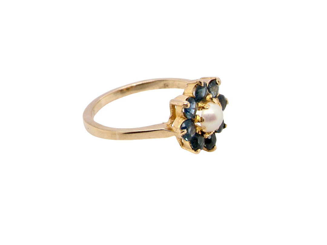 A yellow gold ring featuring a flower-shaped setting with dark blue gemstones and a central white pearl, shown on a plain white background.