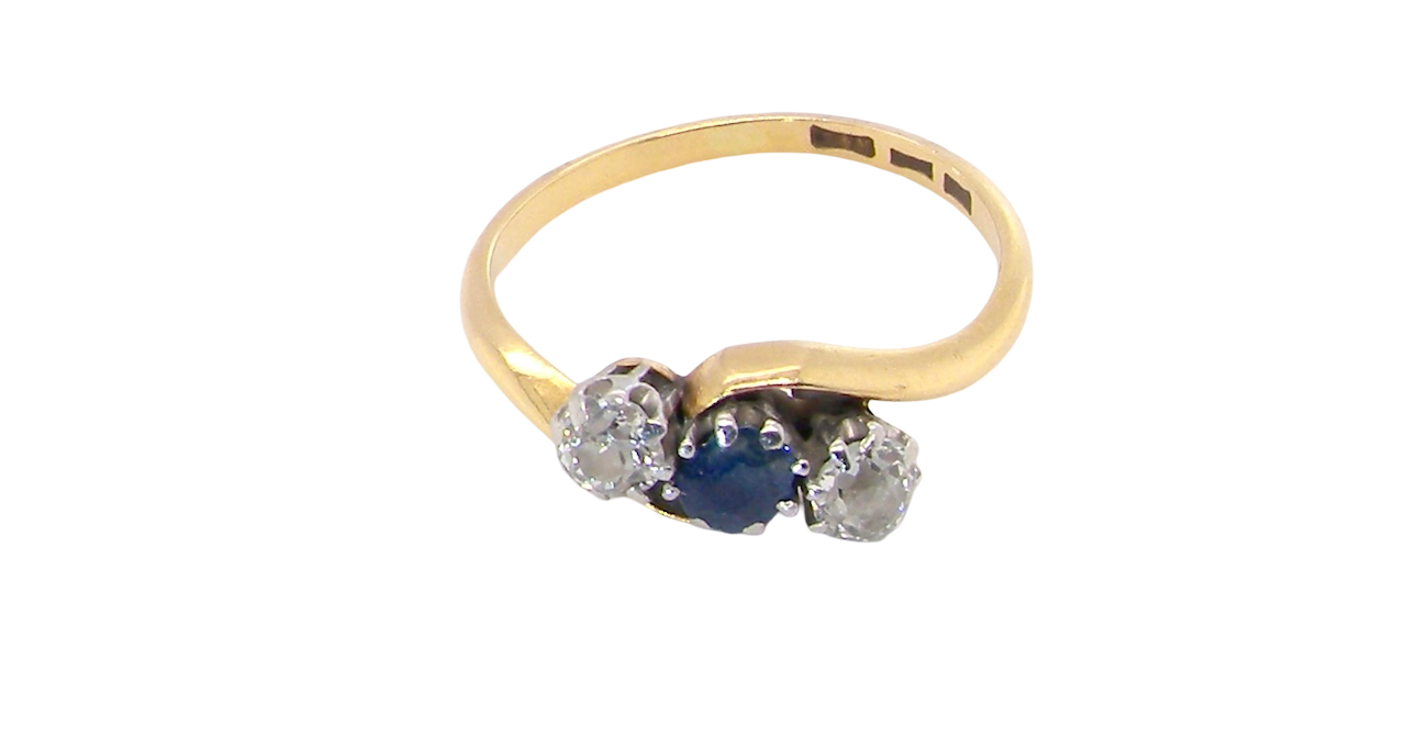 top An 18 carat gold three stone cross over ring.
Centre stone a round sapphire 4mm diameter. Each side a brilliant cut white diamond 4mm diameter (0.25 carats estimated), colour i-k, clarity Si.