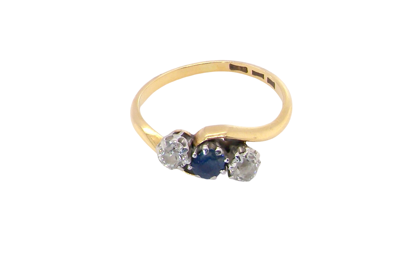 top An 18 carat gold three stone cross over ring.
Centre stone a round sapphire 4mm diameter. Each side a brilliant cut white diamond 4mm diameter (0.25 carats estimated), colour i-k, clarity Si.