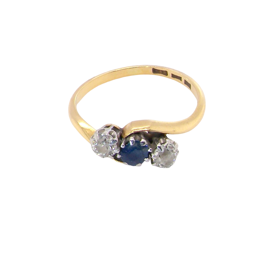 top An 18 carat gold three stone cross over ring.
Centre stone a round sapphire 4mm diameter. Each side a brilliant cut white diamond 4mm diameter (0.25 carats estimated), colour i-k, clarity Si.