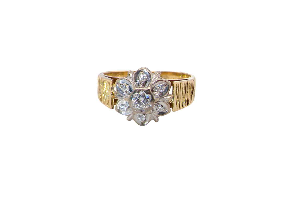 typical design from the 1960s/1970s era, a daisy cluster set with brilliant cut white diamonds and and a bark design finish to both shoulders.
Central diamond measures 2.5mm, and an outer halo of six more diamonds, at 1.2mm each and illusion set in white gold. (Total est carat weight 0.15 carats). Head measures 10mm. The band and shoulders test as 18 carat gold. 