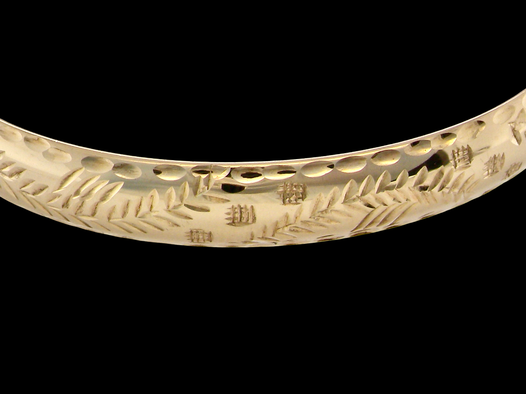 A close-up of a polished yellow gold bangle bracelet featuring engraved leaf and branch motifs, set against a solid black background.