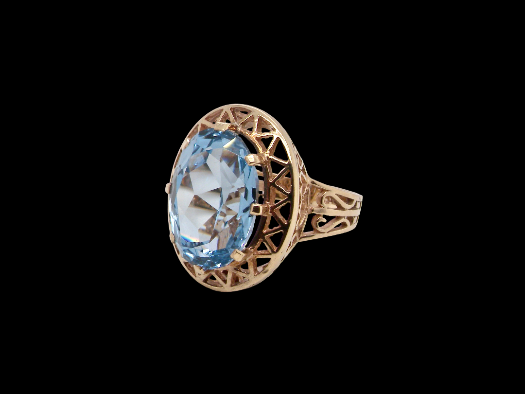 An antique-style gold ring featuring a large oval-cut light blue gemstone, set in an openwork filigree band, shown against a solid black background.
