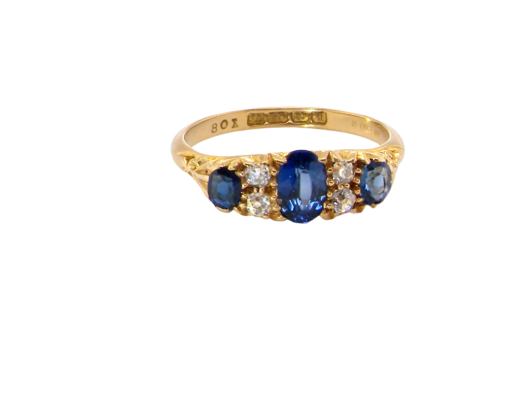 A yellow gold ring featuring a central oval blue sapphire flanked by round and oval blue sapphires and small clear diamonds, shown on a plain white background.