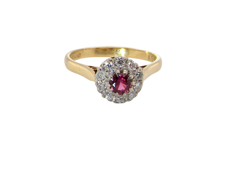 A yellow gold ring featuring a central round pinkish-red ruby gemstone surrounded by a halo of small clear stones, set against a plain white background.