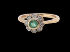 A yellow gold ring featuring a central round green emerald-cut gemstone surrounded by a halo of small clear stones, set on a solid black background.
