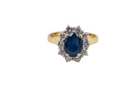 A yellow gold ring featuring an oval-cut dark blue sapphire center stone surrounded by a halo of small clear stones, set against a plain white background.