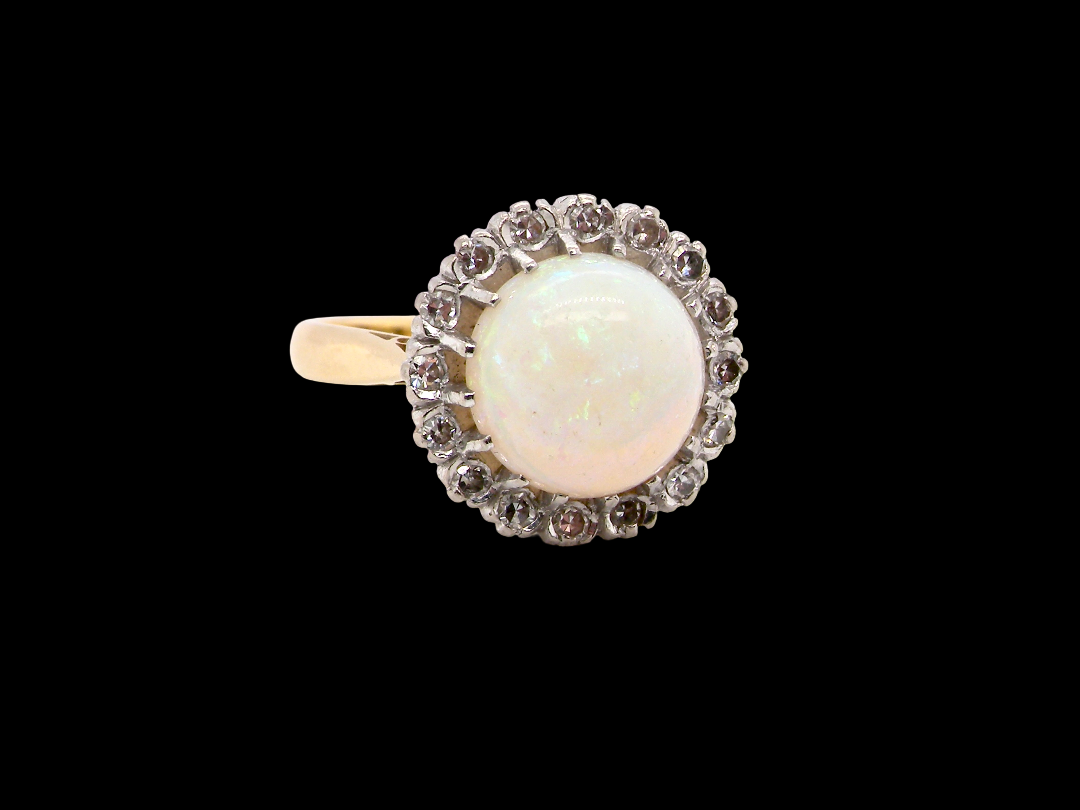 A vintage-style ring featuring a round white opal center stone surrounded by a halo of small clear stones, set on a yellow gold band, photographed against a solid black background.