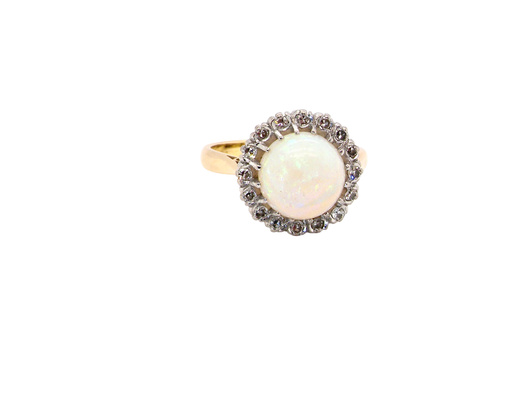 A vintage-style ring featuring a round white opal center stone surrounded by a halo of small dark stones, set on a yellow gold band, isolated on a white background.