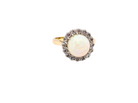 A vintage-style ring featuring a round white opal center stone surrounded by a halo of small dark stones, set on a yellow gold band, isolated on a white background.
