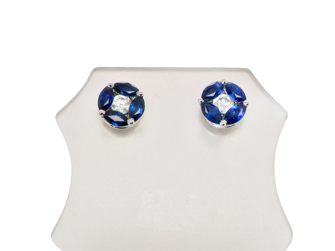 A pair of round stud earrings with deep blue gemstones arranged in a flower shape around a clear central stone, set in silver-toned metal, displayed on a white stand against a white background.
