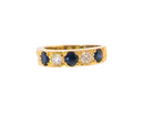 A yellow gold eternity-style ring featuring alternating round dark blue sapphires and clear round diamonds, shown against a plain white background.