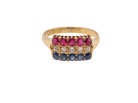 A yellow gold ring featuring three rows of round-cut gemstones: a top row of vibrant pink rubies, a middle row of clear diamonds, and a bottom row of deep blue sapphires, set against a plain white background.