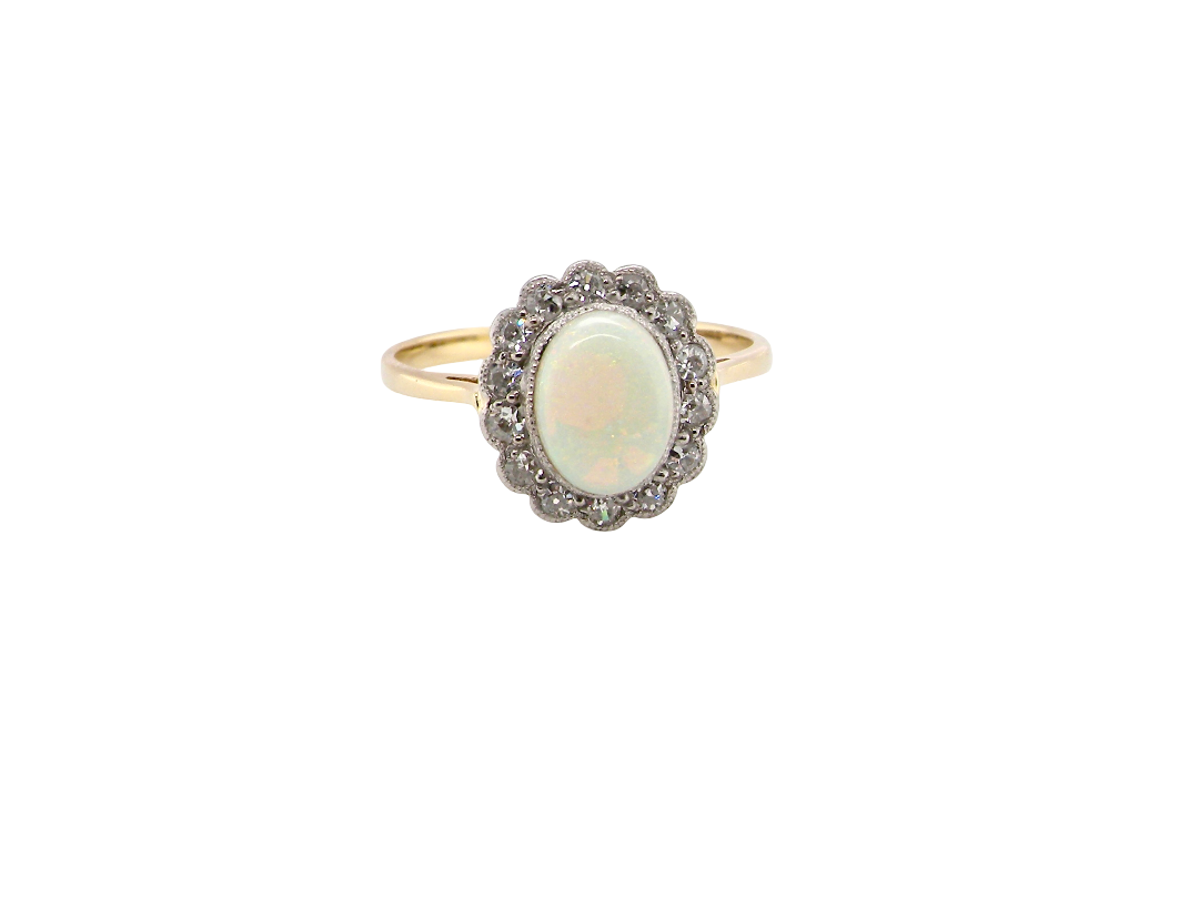 A vintage-style ring featuring an oval cabochon opal surrounded by a halo of small clear stones, set on a yellow gold band, isolated on a white background.