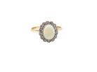 A vintage-style ring featuring an oval cabochon opal surrounded by a halo of small clear stones, set on a yellow gold band, isolated on a white background.