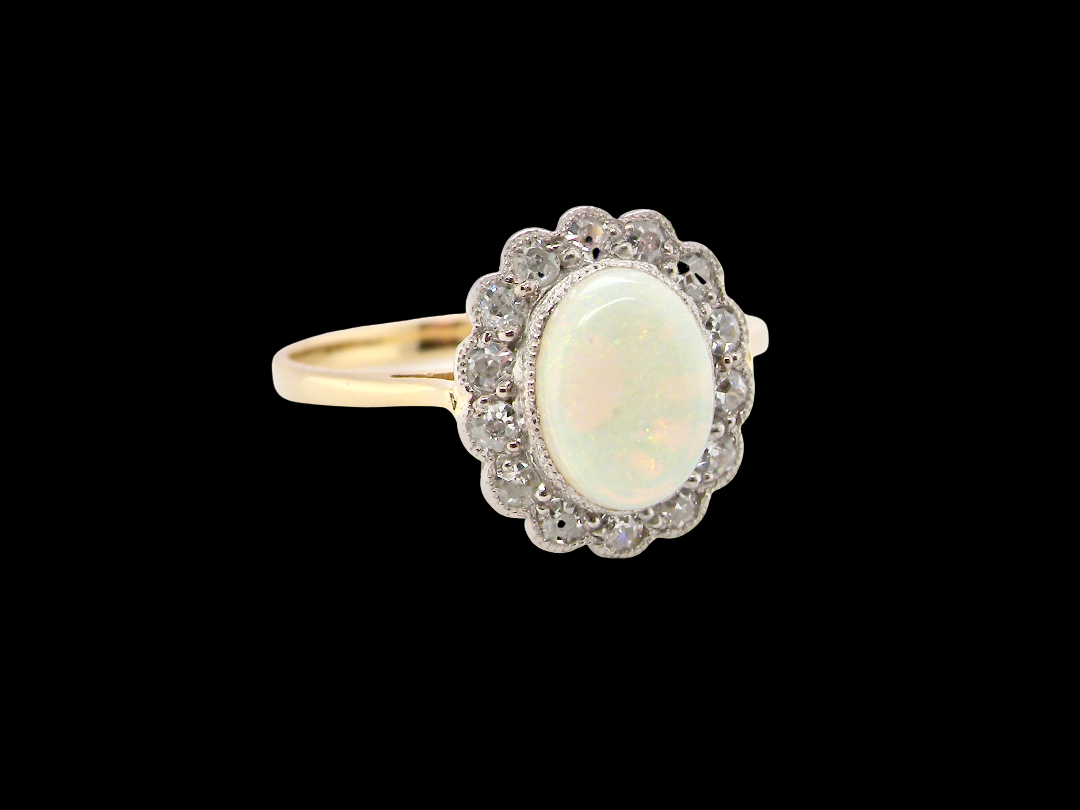 A yellow gold ring featuring an oval white opal center stone surrounded by a halo of small clear stones, set against a solid black background.