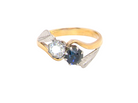 A two-tone ring featuring a yellow gold band, a silver-toned leaf-shaped setting, a clear round-cut gemstone, and a dark blue oval-cut gemstone, shown on a plain white background.
