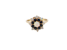 A gold ring featuring a flower-shaped cluster with a central round iridescent opal, surrounded by dark blue sapphires and smaller white opals, set against a plain white background.