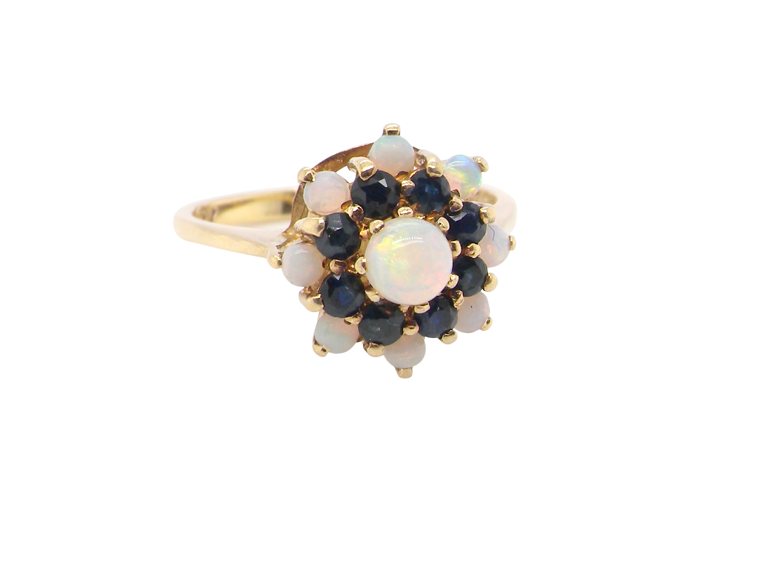 A gold ring featuring a flower-shaped cluster with a central oval white opal surrounded by dark blue sapphires and smaller round white opals, set against a plain white background.