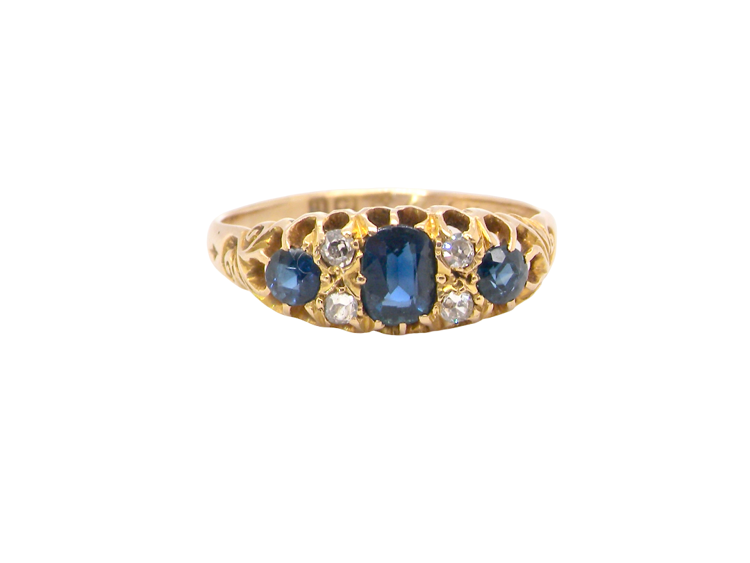 A vintage-style yellow gold ring featuring a central oval blue sapphire flanked by smaller round blue sapphires and clear round diamonds, set against a plain white background.