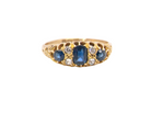 A vintage-style yellow gold ring featuring a central oval blue sapphire flanked by smaller round blue sapphires and clear round diamonds, set against a plain white background.